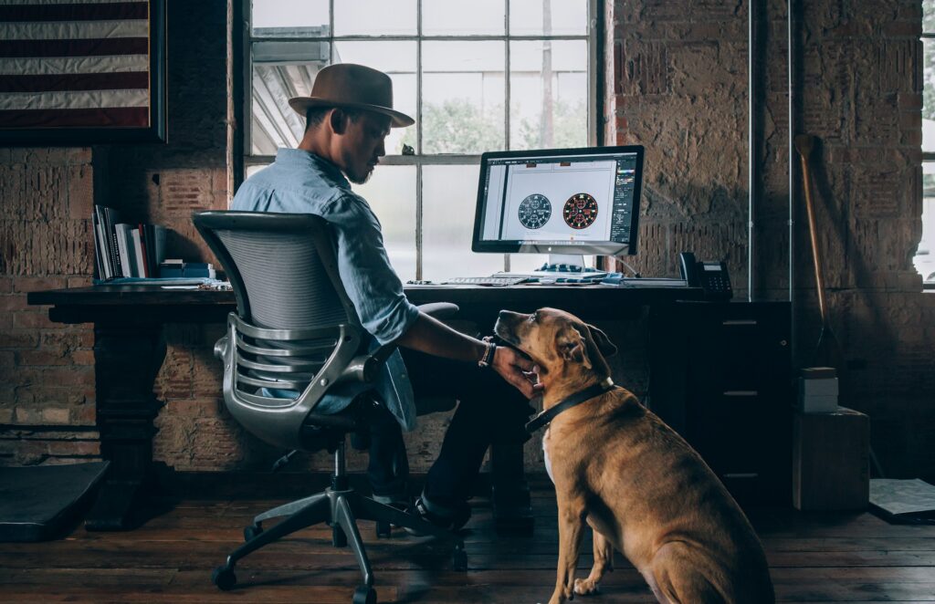 デスクに向かうハットを被った男性の横に犬がいる